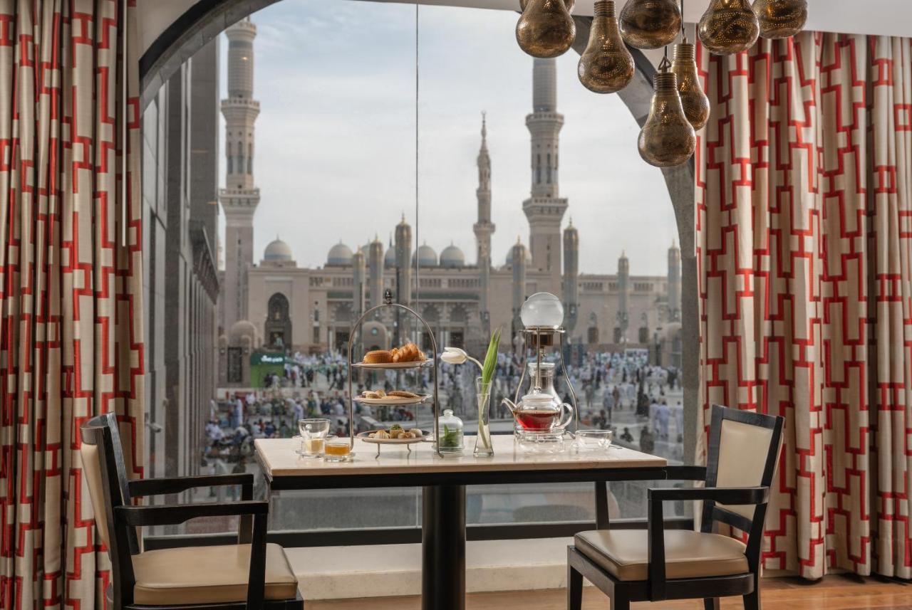 فندق شهد المدينة بإدارة اكور المدينة المنورة المظهر الخارجي الصورة The photo shows a dining setup inside a room with a large window. A table is set for a meal, featuring a tiered stand with various foods, a teapot, and cups. The room is decorated with elegant furniture and patterned curtains. Through the window, the