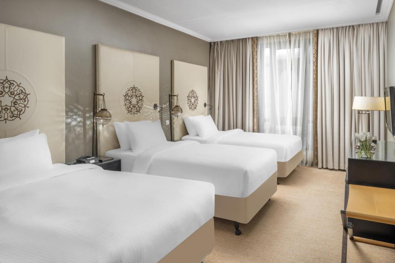 فندق شهد المدينة بإدارة اكور المدينة المنورة المظهر الخارجي الصورة The photo shows a hotel room featuring three beds, each made with white bedding. The beds are positioned against a light-colored wall adorned with decorative headboards. There are two bedside tables with lamps next to the beds. The room is well-lit, 