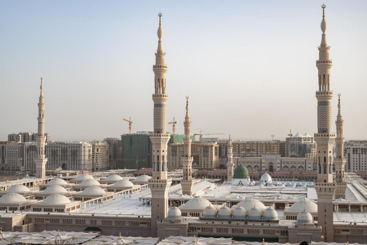 فندق شهد المدينة بإدارة اكور المدينة المنورة المظهر الخارجي الصورة The photo depicts a view of an Islamic mosque complex with several prominent minarets. The architectural style features a blend of intricate designs and large, domed roofs. The scene appears to be set in a modern urban environment, with construction 