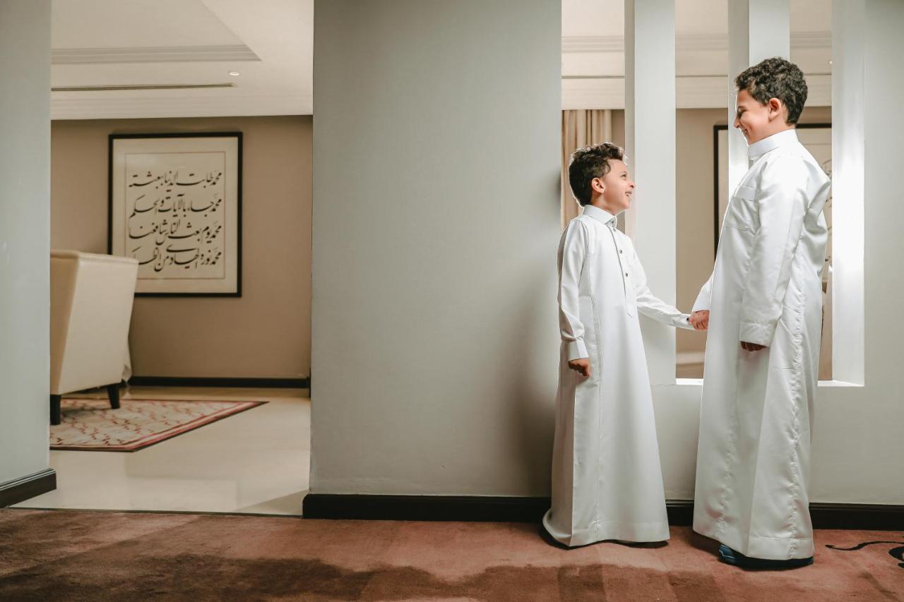 فندق شهد المدينة بإدارة اكور المدينة المنورة المظهر الخارجي الصورة The photo shows two young boys dressed in traditional white clothing, likely thobes. They are standing in a hallway, engaged in a conversation or interaction. The setting appears to be an indoor space with modern decor, including a piece of artwork o