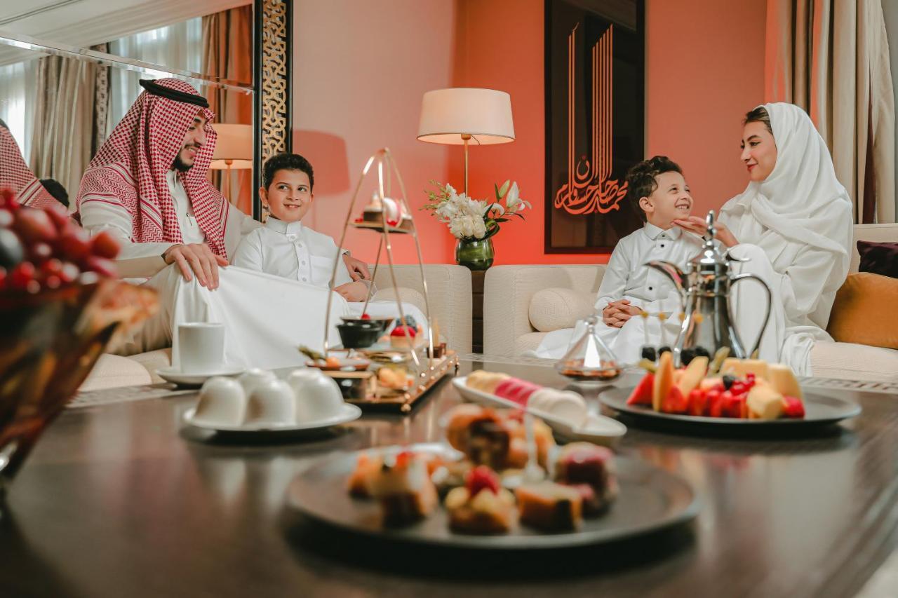 فندق شهد المدينة بإدارة اكور المدينة المنورة المظهر الخارجي الصورة The photo depicts a cozy and inviting indoor setting where a family appears to be spending time together. In the foreground, there are plates of desserts and fruits arranged on a table, showcasing a variety of colorful treats. 

In the background, a 