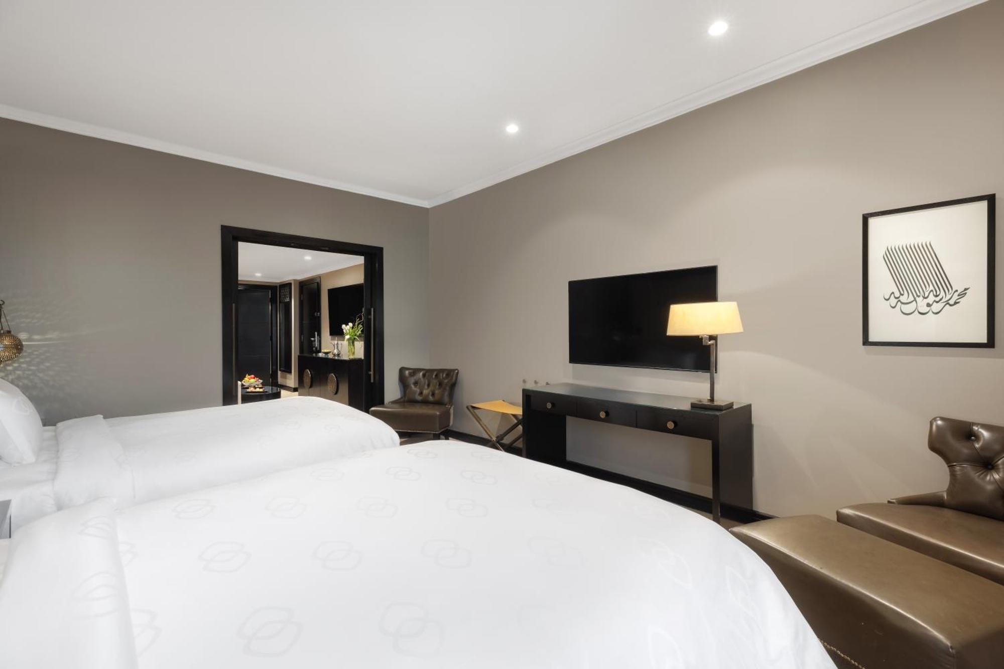 فندق شهد المدينة بإدارة اكور المدينة المنورة المظهر الخارجي الصورة The photo shows a modern hotel room with a neutral color palette. It features two beds with white linens, a stylish lamp on a side table, and a flat-screen TV mounted on the wall. There is a dark wood console table beneath the TV, and a comfortable a
