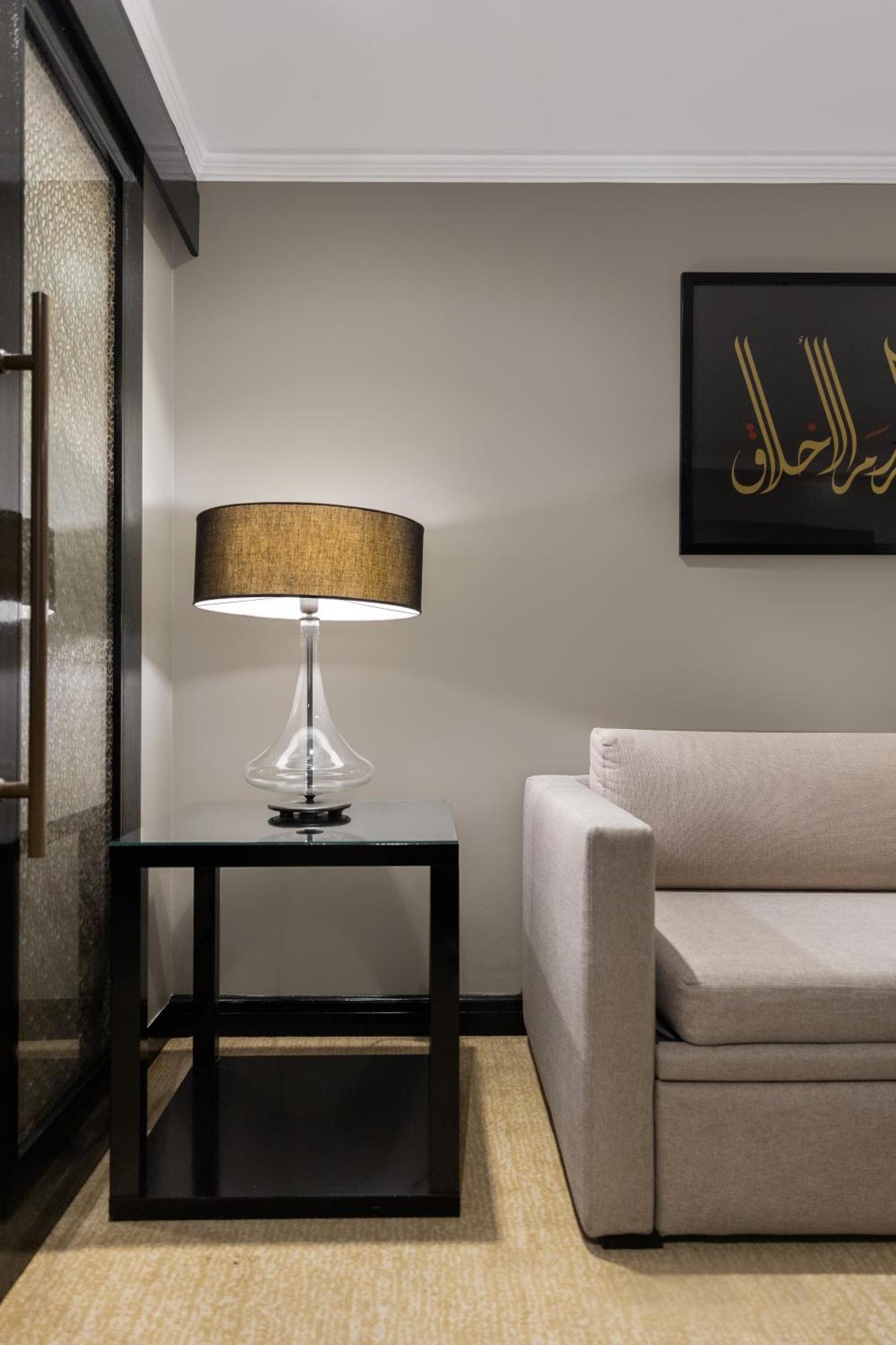 فندق شهد المدينة بإدارة اكور المدينة المنورة المظهر الخارجي الصورة The photo depicts a modern interior setting. On the left, there is a glass table lamp with a cylindrical body and a round, dark-colored lampshade. The lamp is placed on a sleek black side table. To the right, there’s a light-colored sofa with a minim