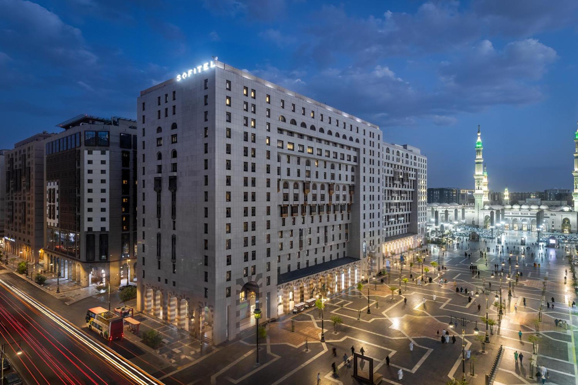 فندق شهد المدينة بإدارة اكور المدينة المنورة المظهر الخارجي الصورة The photo shows a large modern building, likely a hotel, situated in a cityscape at dusk. The structure has a distinct architectural style with multiple stories and illuminated windows. Surrounding the building is a plaza or open area with trees and 
