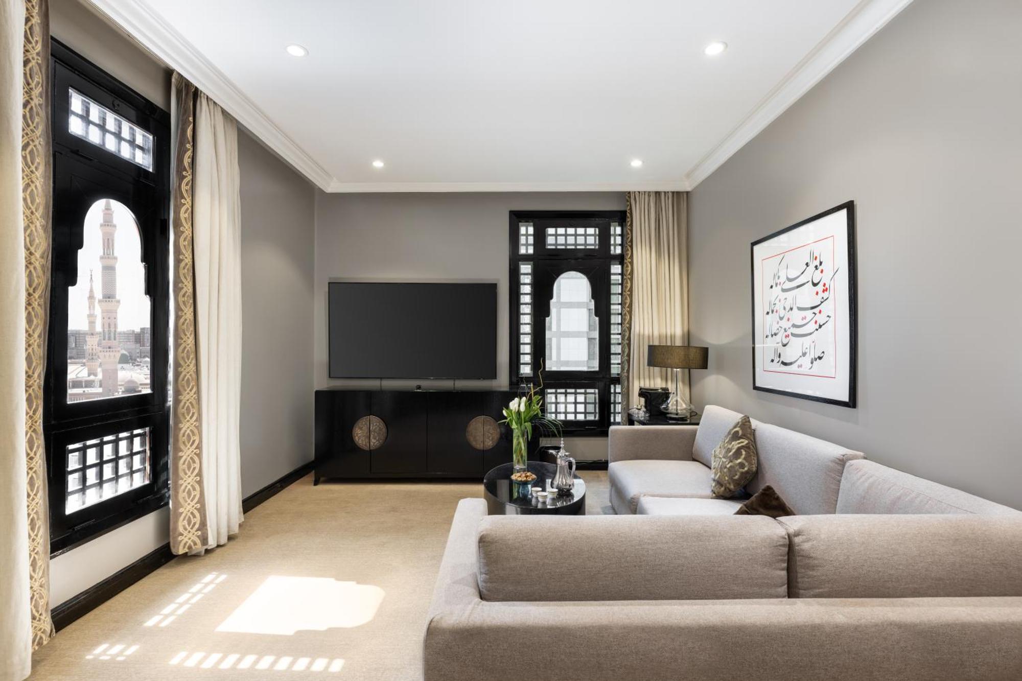 فندق شهد المدينة بإدارة اكور المدينة المنورة المظهر الخارجي الصورة The photo depicts a modern living room with a neutral color palette. There's a comfortable light gray sofa with decorative cushions positioned toward the center. Opposite the sofa, a sleek black TV unit supports a flat-screen television. Large window
