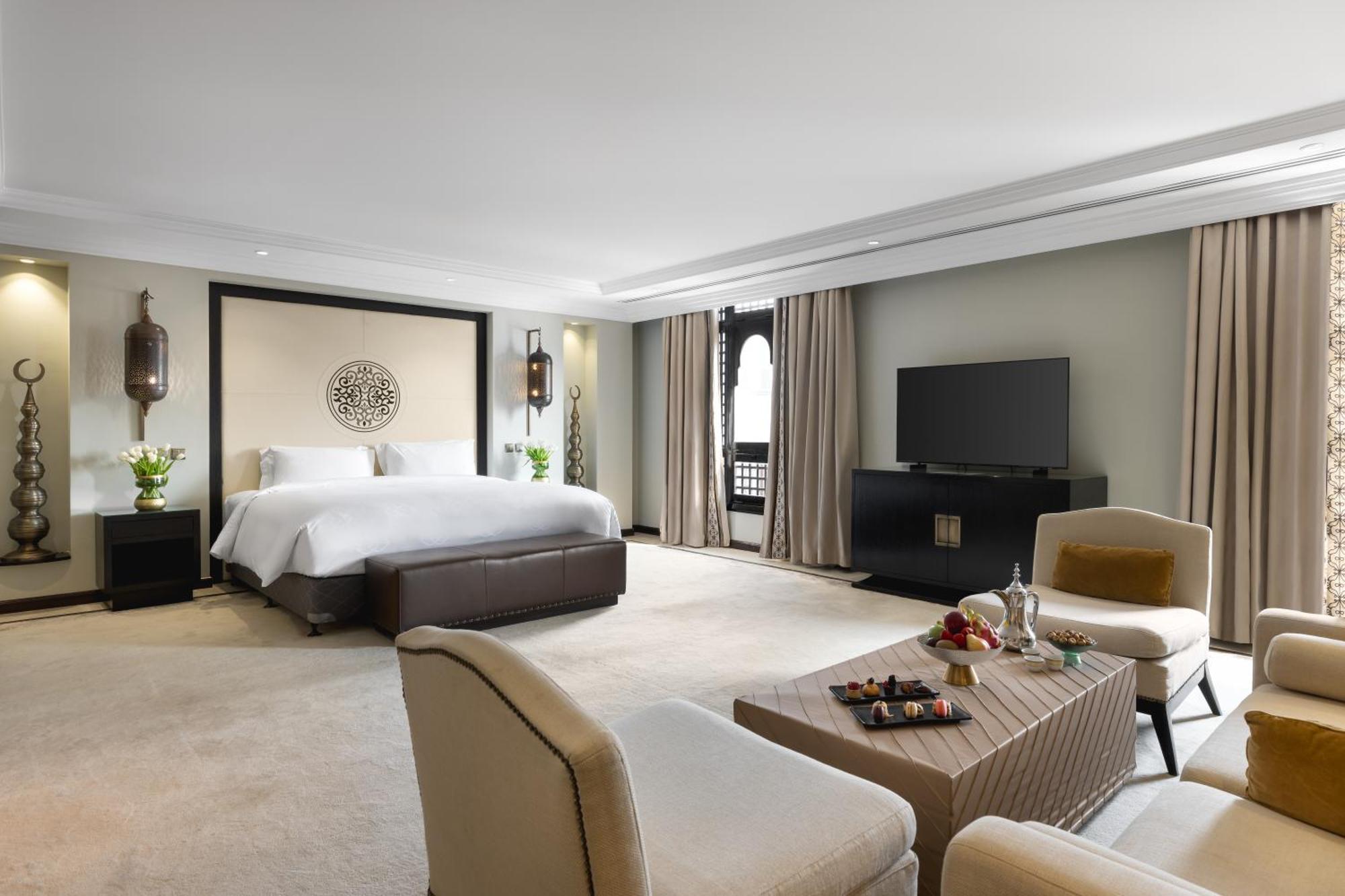 فندق شهد المدينة بإدارة اكور المدينة المنورة المظهر الخارجي الصورة The photo shows a spacious and stylish hotel room. The room features a large bed with a dark headboard, complemented by light-colored bedding. There are two comfortable armchairs facing a coffee table, which holds some snacks and a drink. A modern te