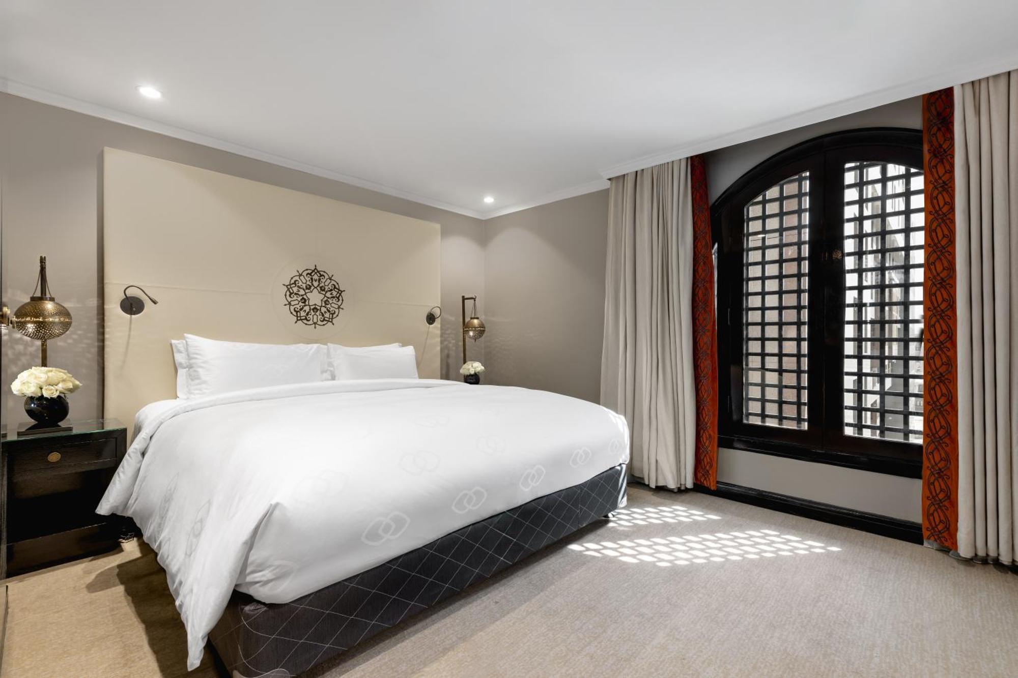 فندق شهد المدينة بإدارة اكور المدينة المنورة المظهر الخارجي الصورة The photo shows a stylish hotel room featuring a large bed with white bedding positioned against a light-colored wall. Decorative elements include a circular wall piece above the bed and bedside lamps. The room has large windows with a unique grid pa