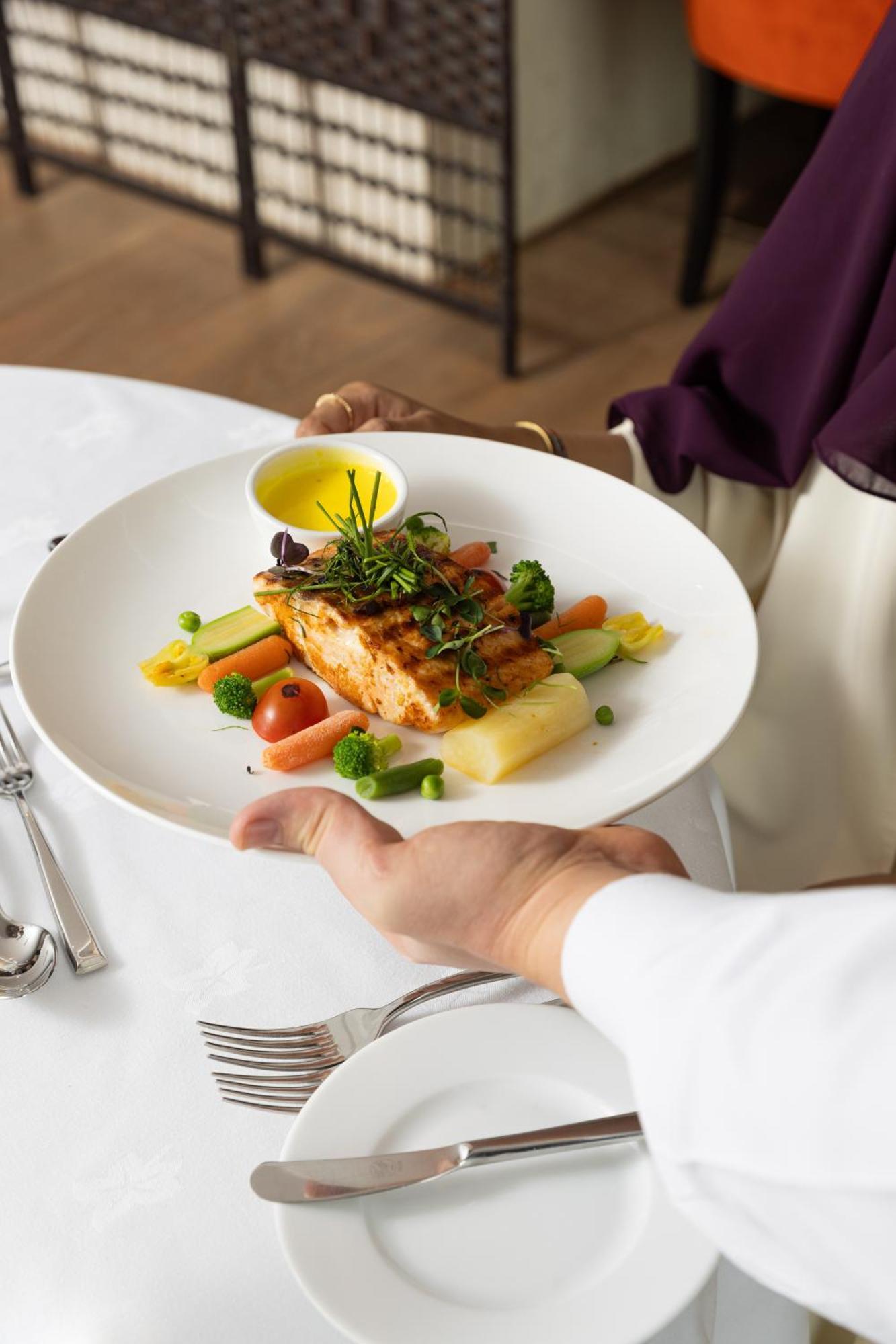 فندق شهد المدينة بإدارة اكور المدينة المنورة المظهر الخارجي الصورة The photo shows a plate of food being presented. The dish features a piece of grilled salmon on a bed of vegetables, including small tomatoes, green peas, and other colorful vegetables. There is also a drizzle of yellow sauce, likely a dressing or oi
