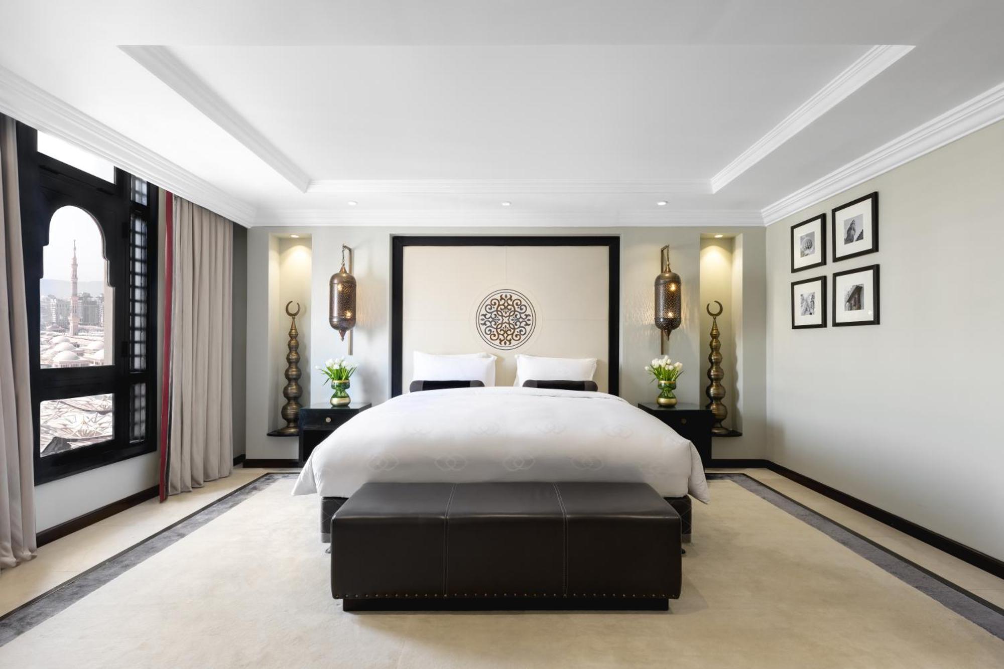 فندق شهد المدينة بإدارة اكور المدينة المنورة المظهر الخارجي الصورة The photo shows a modern and stylish bedroom. It features a large bed with white linens, placed against a decorative wall with a circular design. On either side of the bed, there are elegant wall lamps illuminating the space. The room has a soft colo