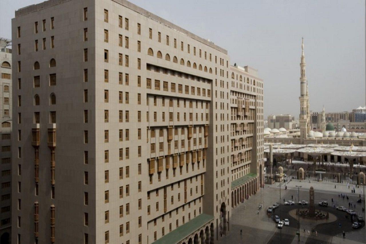 فندق شهد المدينة بإدارة اكور المدينة المنورة المظهر الخارجي الصورة The photo shows a large modern building with a facade that features numerous windows and architectural detailing. It is situated in an urban setting, with another prominent structure, likely a mosque or minaret, visible in the background. The surroun